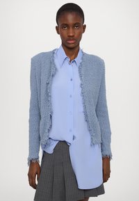Cardigan azzurro chiaro con texture e bordi sfrangiati, indossato sopra una camicia azzurro chiaro con bottoni, abbinato a una gonna plissettata grigia.