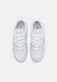 Baltas ādas sporta apavi ar gaiši ziliem akcentiem un perforētu dizainu uz pirksta. Iezīmē zelta "LACOSTE" logo un plakani baltie šņores.