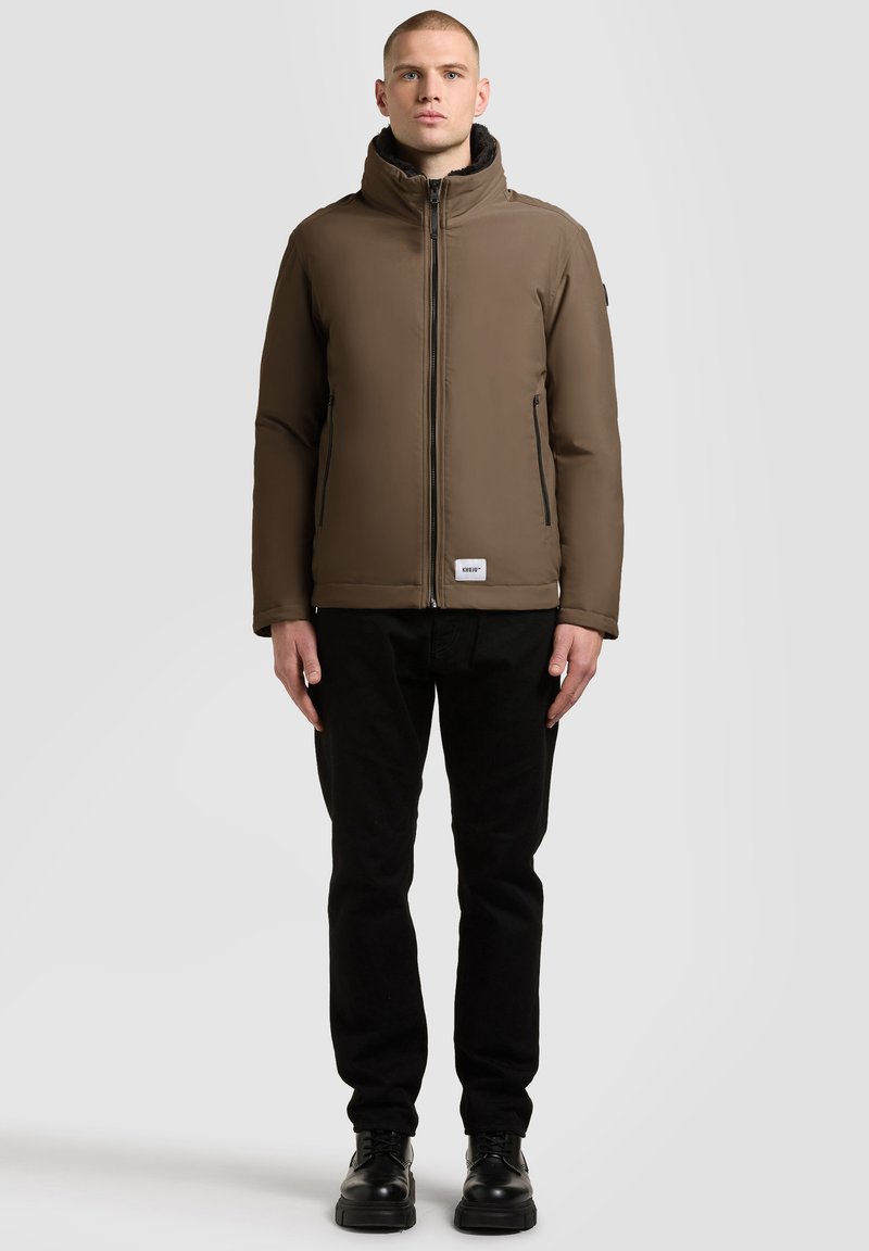 Brown insulated jacket with high collar, zip closure, front pockets, and a logo patch. Worn with black pants and black shoes.