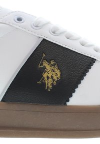 Sneaker bianca e nera con pannello in pelle texturizzata, caratterizzata da un logo d'oro di un giocatore di polo sul lato e una suola in gomma marrone.