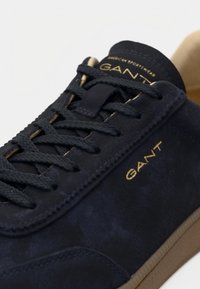 Tmavomodré semišové tenisky s okrúhlou špičkou, plochou gumovou podrážkou, zlatými detailmi loga a šnurovacím dizajnom s tmavými štruktúrovanými šnúrkami.