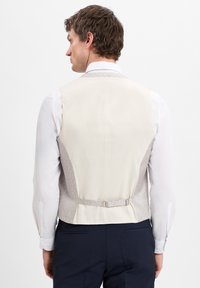 Beige Weste mit strukturiertem grauem Rücken, schlanker Schnitt, verstellbarer Schnalle, getragen über einem weißen Hemd mit spitzem Kragen und Knopfmanschetten.