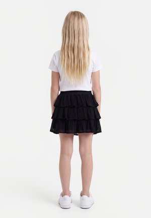 Kind mit langen blonden Haaren, steht mit dem Rücken zur Kamera, trägt ein weißes T-Shirt, einen schwarzen Stufenrock und weiße Turnschuhe vor weißem Hintergrund.