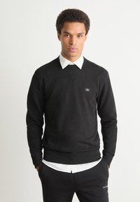 Sweater de algodão preto com mangas longas, decote redondo, punhos canelados e um pequeno logotipo no peito, sobreposto a uma camisa branca com colarinho.