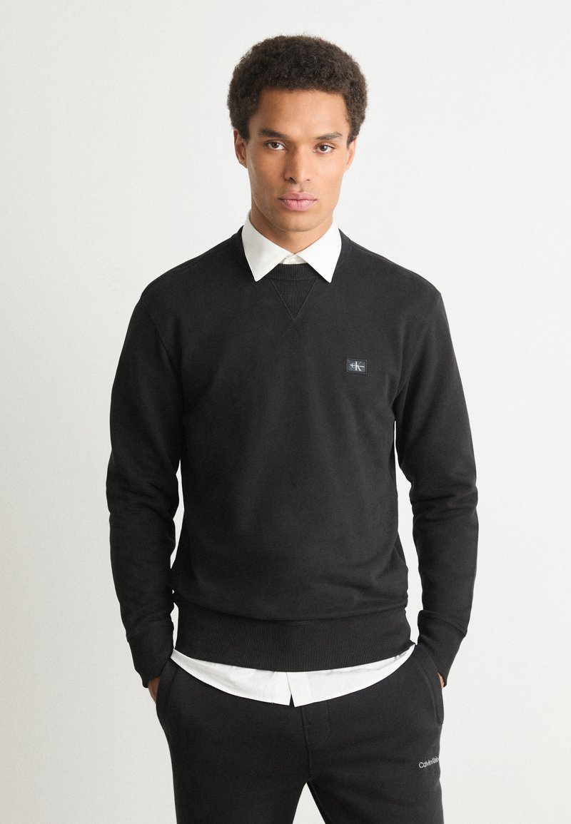 Sweater de algodão preto com mangas longas, decote redondo, punhos canelados e um pequeno logotipo no peito, sobreposto a uma camisa branca com colarinho.