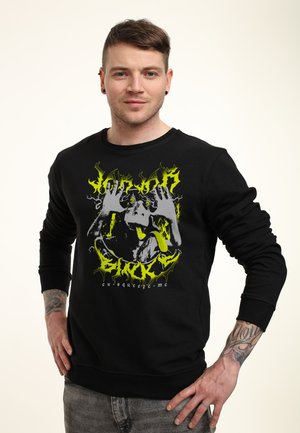 Młody mężczyzna z krótkimi brązowymi włosami, ubrany w czarną bluzę z neonowo-żółtą grafiką małpy robiącej minę i tekstem, stojący z rękami na biodrach.