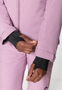 Heled lilla kahekihiline jope mustade mansettidega, millel on lukuga esiosa, velcro reguleerimine mansettidel ja trükitud logo detail varrukal.