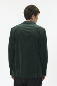 Vyriškis, stovintis atsuktas nugara, dėvi tamsiai žalią aksominį švarką ir juodus kelnes, priešais baltą, paprastą foną.