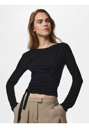 Donna con lunghi capelli castani che indossa una maglia nera a maniche lunghe stile incrociato e pantaloni beige plissettati a vita alta con le mani nelle tasche.