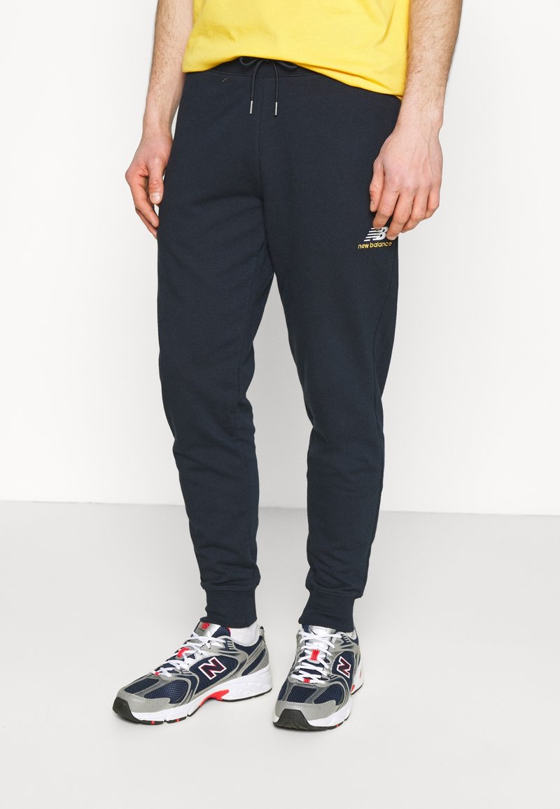 Navyblauwe sweatpants met een taps toelopende vorm, een trekkoord in de taille en een klein gouden New Balance-logo. Gecombineerd met grijze en navy sport schoenen.
