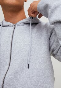 Lys grå zip-up hoodie lavet af blødt materiale, med en hætte med snøre, ribbede manchetter og en glat lynlås med sorte detaljer.