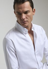 Camisa blanca de botón, hecha de algodón suave, con cuello. En la parte delantera presenta un detalle de logo en el lado izquierdo del pecho.