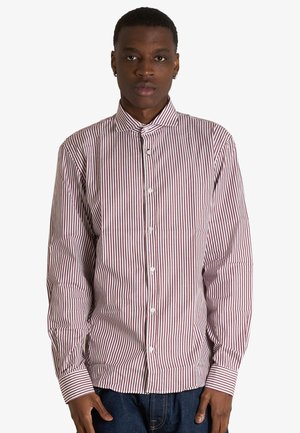 Uomo che indossa una camicia a maniche lunghe abbottonata con righe verticali rosse e bianche, in piedi davanti a uno sfondo bianco semplice.