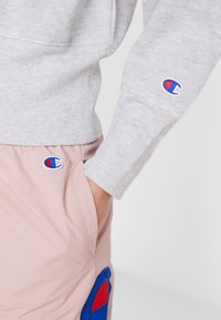 Sweatshirt gris avec des poignets côtelés, associé à un short rose clair avec un patch de logo bleu et rouge. Vue rapprochée mettant en avant le tissu et les logos.