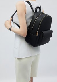 Musta nahkareppu, jonka muoto on jämäkkä, kultaiset vetoketjut, yläkahva ja etutasku. Keskellä selkää valkoisen paidan päällä.