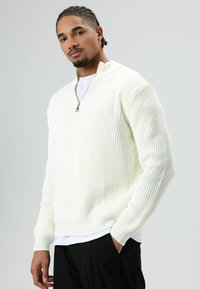 Man with braided hair wearing a white ribbed half-zip sweater, white shirt underneath, and black pants with hand in pocket.