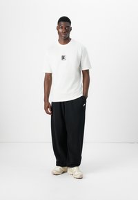 Hvid T-shirt med et sort grafisk logo, parret med løse sorte bukser og lyse sneakers. Glat stof, afslappet pasform, casual design.