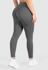 Persona vestida con leggings de talle alto en gris oscuro y zapatillas blancas, de pie con una mano relajada a su lado frente a un fondo sencillo.