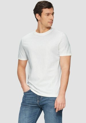 Mann mit kurzen dunklen Haaren, der ein einfaches weißes T-Shirt mit kurzen Ärmeln und blaue Jeans trägt, mit einer Hand in der Tasche stehend und nach rechts schauend.