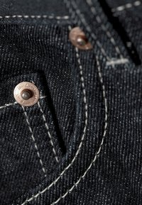 Tmavě modrý denimový materiál s měděnými nýty a kontrastním béžovým prošíváním. Detailní záběr ukazuje kapsu a strukturu materiálu.
