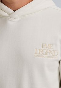 Biela mikina s kapucňou vyrobená z mäkkého textúrovaného materiálu. Na hrudi sa nachádza béžový vyšitý nápis: "PME LEGEND EXPLORING THE ELEMENTS."