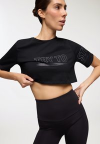 Maglietta nera cropped con la frase "TRY TO" in un bold grigio, abbinata a leggings neri a vita alta. Tessuto morbido e liscio.