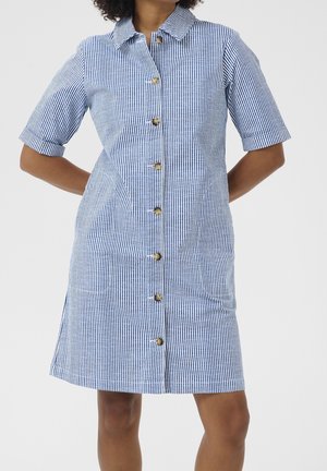 Kobieta w niebiesko-białej sukience w pionowe paski z krótkim rękawem, kołnierzykiem i zapięciem na guziki, stoi z rękami za plecami.