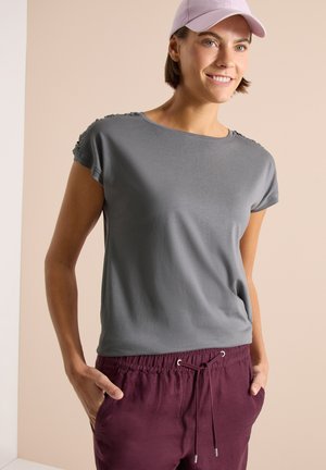 Kvinde smilende iført en lyse lilla kasket, grå kortærmet skjorte og bordeaux bukser med snøre, med hænderne i lommerne foran en beige baggrund.