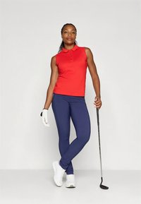 Polo vermelho sem mangas com colarinho, combinado com leggings azul-marinho e ténis brancos. Segurando um taco de golfe, usando uma luva branca de golfe.