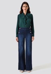 Blouse verte avec un col à nœud, associée à un jean bleu foncé taille haute à jambes larges, doté de boutons dorés et d'une texture en denim lisse.