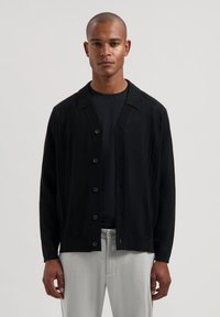 Cardigan noir en maille avec un design texturé, fermeture à boutons et col. Porté sur une chemise noire et associé à un pantalon gris clair.