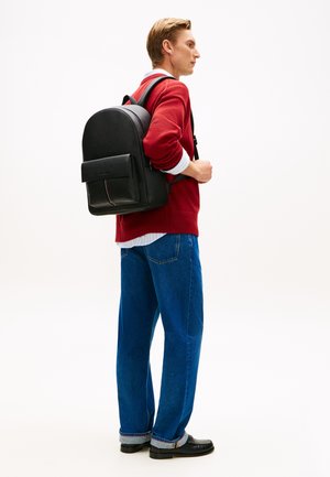 Sac à dos en cuir noir avec une surface texturée, profil mince, poche avant avec rabat, et un accent rose subtil. Porté par une personne en tenue décontractée.