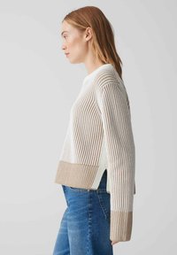 Gestreifter Strickpullover in Weiß und Beige. Kürzes Design mit weiten Ärmeln, Seitenschlitzen und gerippter Textur. Kombiniert mit Jeans.
