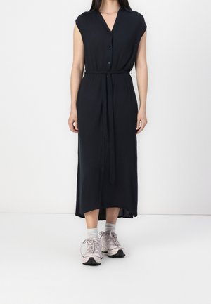 Frau trägt ein ärmelloses schwarzes Midikleid mit Knöpfen und Taillengürtel sowie hellrosa Sneakers mit weißen Socken vor weißem Hintergrund.
