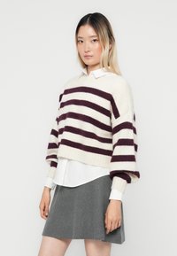 Gestreepte trui in crème en bordeaux, zachte brei, cropped ontwerp, met verlaagde schouders en textiel. Gelaagd over een witte blouse.