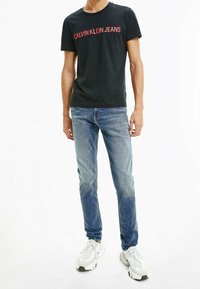 Černé bavlněné tričko s potiskem "CALVIN KLEIN JEANS" v červené barvě. Nosené s světle modrými džínami a bílými teniskami. Jednoduchý design s kulatým výstřihem.