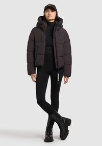 Pufjakke i mørkegrå, med hætte, frontlynlås og quiltet tekstur. Parret med sorte leggings og klodsede sorte støvler.