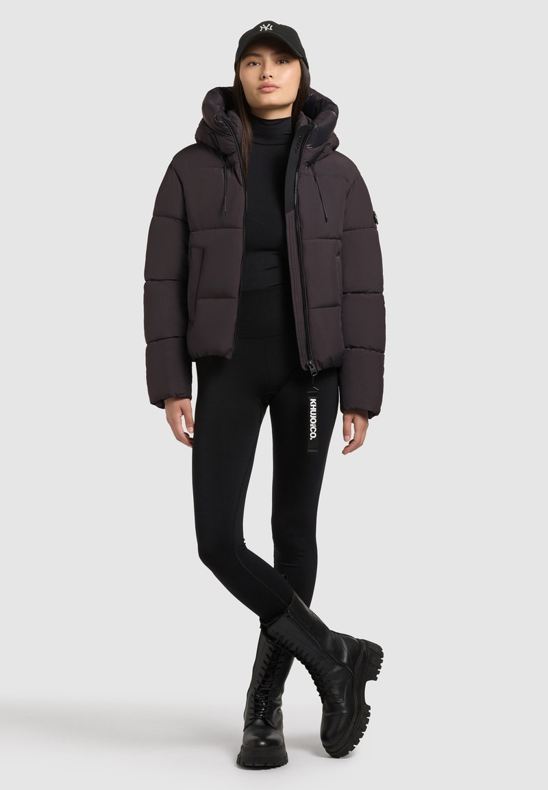 Pufjakke i mørkegrå, med hætte, frontlynlås og quiltet tekstur. Parret med sorte leggings og klodsede sorte støvler.