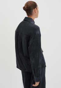 Giacca in velluto a coste scuro con un fit rilassato, caratterizzata da un ampio colletto e polsini con bottoni, che presenta una texture a coste e un design minimalista.