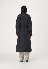 Abrigo largo negro hecho de un material acolchado, con cintura ceñida con cinturón, gran cuello y una textura suave. Se lleva sobre pantalones gris claro.