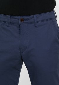 Pantaloni di cotone blu navy con chiusura a bottoni, taglio dritto e cuciture sottili. Presenta una tasca frontale su ciascun lato.