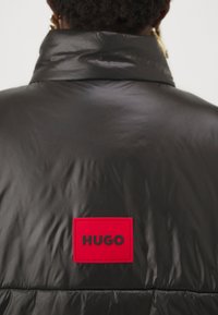 Veste en doudoune noire avec un col haut, une texture brillante et une grande étiquette de logo rouge rectangulaire avec des lettres noires au dos.