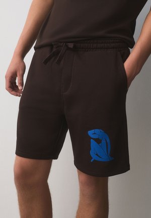 Homme portant un short noir avec taille élastique et motif abstrait bleu de silhouette humaine sur la jambe gauche, mains dans les poches.