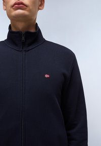 Giacca a zip blu navy con colletto alto, realizzata in tessuto morbido, con una piccola bandiera norvegese ricamata sul lato sinistro del petto.