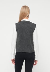 Femeie cu părul lung blond, îmbrăcată cu o cămașă albă, un veston din tricot fără mâneci de culoare gri închis și pantaloni negri, arătată din spate, pe un fundal simplu.