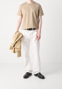 Beige bomulls t-shirt med liten logotyp, vita denimbyxor och en ljusbeige jacka draperad över en arm. Svart bälte och läderskor.