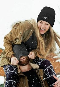 Black knitted beanie with ribbed texture and a white graphic patch. Wearing a warm olive parka over a dark top; snow falling in the background.