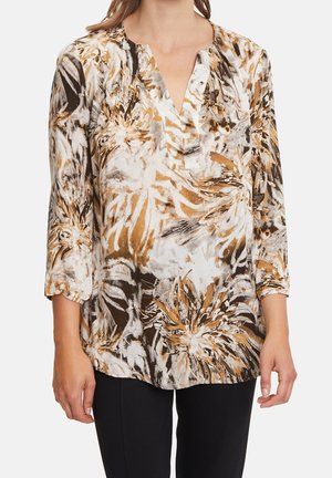 Kobieta ubrana w bluzkę z długim rękawem o abstrakcyjnym wzorze kwiatowym w kolorach brązowym, czarnym i białym, w połączeniu z czarnymi spodniami.