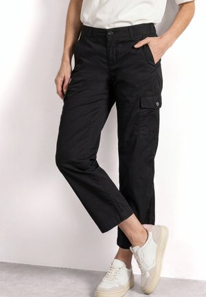 Person trägt schwarze Cargohosen und weiße Sneakers mit beigen Akzenten und steht vor einer hellen Wand, die Hände teilweise in den Taschen.