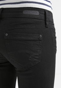 Jeans de mezclilla negra con un diseño ajustado, bolsillos traseros con cierres de botón y patrones de costura sutiles. Etiqueta de marca minimalista visible.